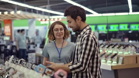 In-the-electronics-store-female-consultant-gives-professional-advice-to-a-young-man.-He-considers-to-buy-a-new-smartphone-and-needs-expert-opinion.-Store-is-modern,-bright-and-has-all-the-new-devices.-Blurred-background.-Slow-motion