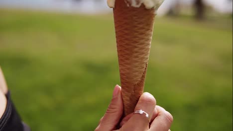 Close-up-cute-brunette-sensual-girl-licking-ice-cream-on-a-cone---white-and-yellow-scoops.-While-looking-into-camera-and-smiling.-Captured-in-side-view.-Green-outdoors