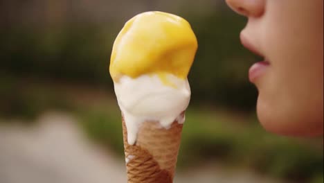 Joyful-brunette-sensual-girl-licking-ice-cream-on-a-cone---white-and-yellow-scoops.-While-looking-into-camera-and-smiling.-Captured-in-side-view.-Green-outdoors.-Close-up