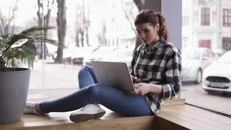 Beautiful-young-brunette-woman-working-on-her-laptop.-Sitting-near-the-window-in-a-cafe-with-modern-interior.-Blurred-street-background