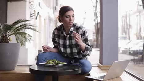Attractive-young-woman-eating-lunch-in-light-cafe,-enjoying-her-salad,-looking-at-laptop.-Front-view