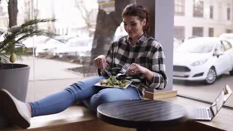 The-girl-sits-leaning-against-the-window.-A-plate-on-her-legs.-She-mixes-the-green-leaves-of-the-salad-with-a-fork-and-knife.-Tastes-the-food-and-enjoys-it.-Slow-motion