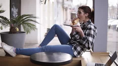 Side-view-of-a-young,-pretty-girl-sitting-in-a-cafe-on-a-wooden-seat-next-to-the-bright-windows.-The-girl-is-eating-a-croissant.-She-chews-and-her-eyes-closed.-Enjoy-the-time