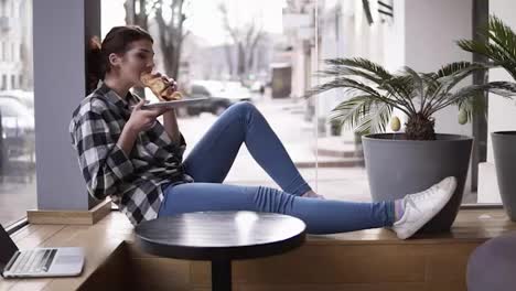 A-brunette-girl-in-a-plaid-shirt-and-jeans-sits-with-outstretched-legs-by-the-window-and-enjoys-her-croissant.-Smiling.-Blurred-street-background