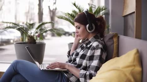 Beautiful,-cheerful-woman-listening-to-music-on-laptop-at-trendy-room-with-headphones-sitting-on-a-couch-in-casual.-Happy,-enjoy-her-time.-Side-view