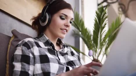 Close-up-footage-of-beautiful,-cheergul,-brunette-girl-in-plaid-shirt.-Happy-girl,-chatting-with-someone-using-a-laptop.-Headphones.-Indoors