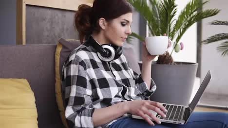 Relaxed,-charming-girl-sits-on-a-sofa-in-the-room,-types-on-a-laptop.-A-big-white-cup-in-her-hand.-The-girl-drinks-cappuccino-and-enjoys-the-time.-Headphones-on-the-neck