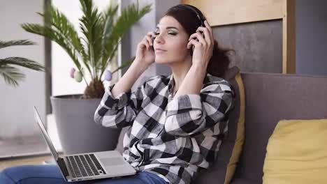 Gorgeous,-young-woman-relaxes-sitting-on-couch,-puts-on-headphones-and-turns-on-music-on-laptop.-Smiles-and-moves-to-the-rhythm-of-music.-Slow-motion