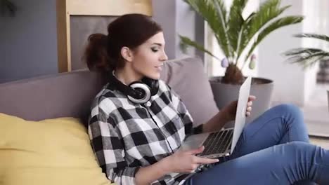 A-trendy-brunette-girl-with-a-laptop-in-her-hands-flops-on-the-sofa-and-starts-typing.-Hedphones-on-neck.-Modern-interior
