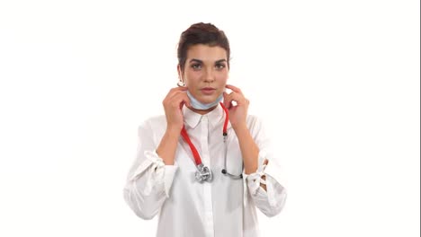 Female-doctor-looking-in-camera-and-taking-on-a-operation-protection-mask.-Close-up-of-a-female-surgeon-with-stethoscope-and-lab-coat-isolated-on-white-background-dressing-up-surgical-mask