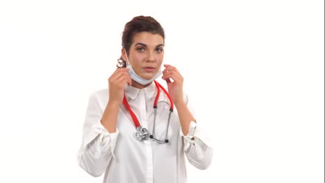 Portrait-of-a-female-doctor-looking-in-camera-and-taking-on-a-protection-mask.-Close-up-of-a-female-surgeon-with-stethoscope-and-lab-coat-isolated-on-white-background-dressing-up-surgical-mask