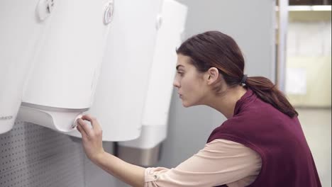 Young-girl-looking-for-water-heater-turns-the-button-on-white-boiler-and-examines-it-in-hardware-store.-Costumer-looking-for-house-appliance.