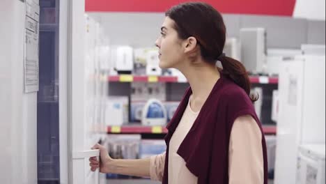 Young-girl-in-her-20's-comes-up-to-a-fridge-in-appliance-store-opens-the-door-and-checks-out-what's-inside.-She-opens-an-upper-plastic-shelf.-Looking-for-household-equipment