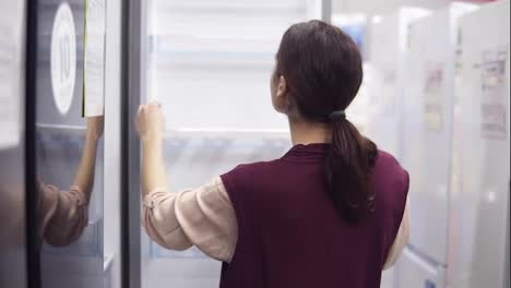Young-brunette-girl-searching-for-perfect-fridge-in-hardware-store.-She-examines-what's-inside-the-white-fridge.-Looking-for-household-equipment