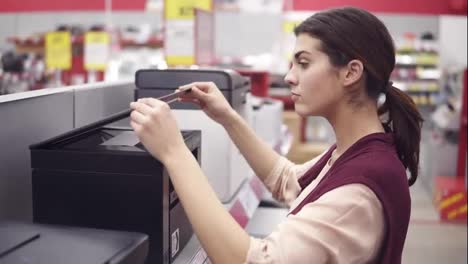 Female-costumer-choosing-new-printer-in-electronics-department-in-appliance-store.-Coming-up-to-a-showcase-row-and-examining-it-carefuly.-Looking-for-household-electronics.