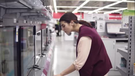Young-female-costumer-choosing-new-TV-set-compares-pricetags-for-different-TV-screens-in-department-of-electronics-in-appliance-store.