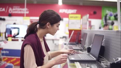 Young-female-costumer-opens-laptop-from-showcase-row-to-examine-it.-Choosing-new-home-electronics.