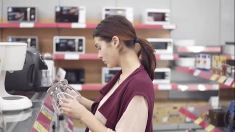 Young-female-costumer-examining-glass-container-for-coffee-mashine-looking-for-perfect-kitchenware-in-appliance-store.-Choosing-new-household-equipment