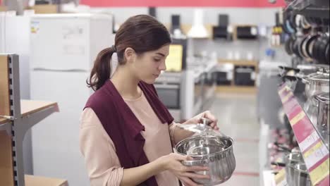 Young-female-costumer-choosing-cook-pot-from-a-variety-of-kitchenware-in-an-appliance-store.-Examining-carefuly-steel-pans-in-a-showcase-row.