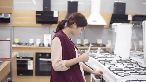 Young-girl-searching-for-perfect-cooktop-in-hardware-store.-She-checks-the-price-turns-burner-buttons-opens-the-oven.-Choosing-household-equipment.-Appliance-store.