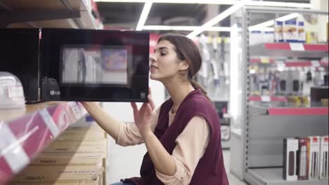 Attractive-girl-in-her-20's-searching-for-microwave-in-appliance-store-opens-black-microwawe-and-checks-out-what-is-inside.-Beautiful-brunette-buying-household-equipment.