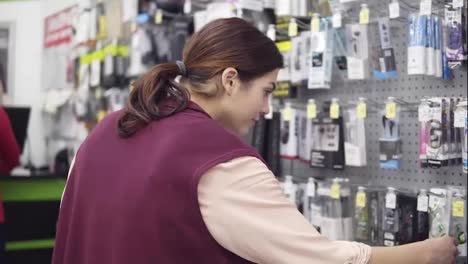 Young-attractive-brunette-girl-trying-to-pick-headphones-reading-labels-on-packagings-on-display-in-electronics-store