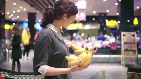 Young-beautiful-brunette-girl-in-her-20's-picking-out-bananas-and-putting-them-into-shopping-cart-at-the-fruit-and-vegetable-aisle-in-a-grocery-store