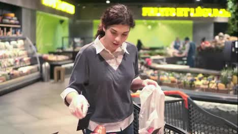 Young-beautiful-brunette-girl-in-her-20's-picking-out-red-apples-into-a-plastic-bag-at-the-fruit-and-vegetable-aisle-in-a-grocery-store.-Frontside-footage.