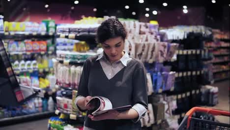 Young-beautiful-brunette-girl-at-her-20's-leafes-through-magazine-and-puts-it-back-on-shelf-trying-to-decide-which-one-to-buy-in-local-supermarket