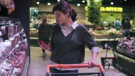 Young-brunette-female-costumer-looking-at-cellphone-while-walking-through-meat-aisle-in-grocery-store.-Girl-takes-salami-stick-from-the-shelf-and-puts-it-into-shopping-cart