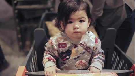 Cute-asian-toddler-boy-is-sitting-in-a-shopping-cart-while-his-mom-is-picking-out-groceries-at-supermarket.