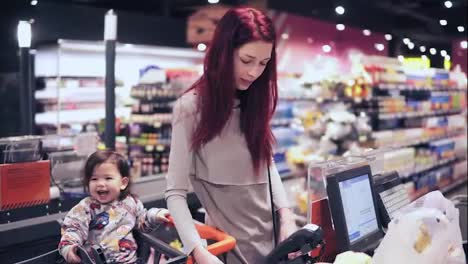 Young-woman-with-red-hair-is-paying-for-purchase-at-checkout-counter-while-her-son-is-laughing,-sitting-in-the-shopping-cart.-Shopping-at-grocery-store.