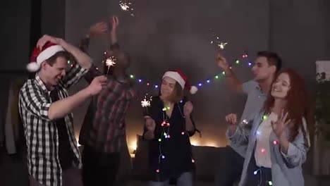 Young-aged-people-enjoying-celebration,-diverse-young-caucasian-friends-hanging-out-during-the-New-Year-Eve---dancing-together-with-bengal-lights-inhands,-having-fun-at-home-decorated-with-garland
