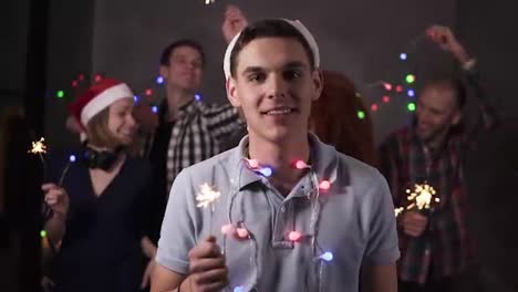 Portrait-of-caucasian-young-guy-with-colorful-lights-on-neck-and-santa-hat-posing-for-camera---smiling,-holding-his-bengal-light-while-his-friends-dancing-and-celebrating-on-the-blurred-background-in-decorated-room.-Friends-celebrating-new-year-together