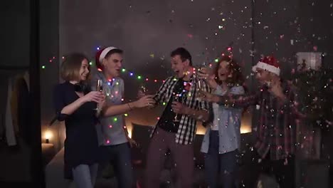 Colorful-celebration-of-Christmas-and-New-year.-Festive-party,-group-of-five-clink-glasses-with-champagne-after-one-guy-explodes-the-confetti-stick.-Happy-celebration-together-with-close-friends-at-home-in-decorated-with-garland-room