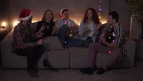 Cheerful-close-friends-celebrating,-having-time-together-at-home.-Happy-friends-sit-around-on-sofa-and-listen-to-guy-singing-and-playing-guitar.-Get-together-at-Christmas-time.-Muffled-light,-garland-lights