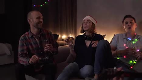 Close-up-footage-of-best-friends-celebrating,-having-time-together-at-home.-Happy-friends-sit-around-on-sofa-and-listen-to-guy-singing-and-playing-guitar.-Singing-together.-Muffled-light,-garland-lights.-Slow-motion