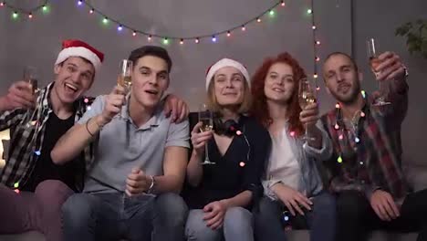 Happy-young-people-drinking-champagne-from-the-glasses-during-celebrate-New-Year-or-Christmas-Eve,-having-great-time-in-relaxing-home-atmosphere-with-garlands-lights-everywhere.-Group-of-young-friends-some-in-Santa's-hats.-Front-view