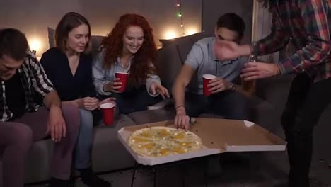 Diverse-people,-friends-got-together-to-eat-pizza-at-home.-Got-delivery-Italian-pizza-with-cheese,-taking-slices,-holding-red-cups-and-bottles.-Starting-home-pizza-party.-Garlands-light-on-the-background