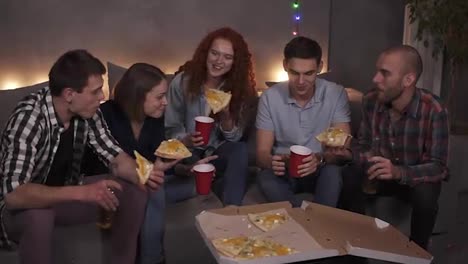 Young-people-cheerful-men-and-women-are-eating-cheesy-pizza,-chatting-relaxing-during-indoor-party-in-apartment-in-a-loft-room.-Delicious-pizza-in-cardbox-is-visible-on-table,-holding-bottles-and-cups-with-drinks