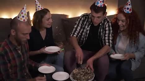 Birthday-guy-in-plaid-shirt-cutting-festive-birthday-cake-in-colorful-cone-hat.-Cheerful-friends-in-cones-enjoying-celebration-in-festive-decorated-room.-Sharing-plate-for-cake-and-smiling