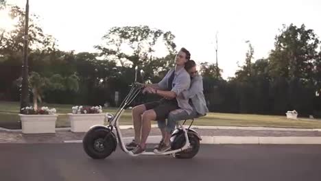 Confident-man-in-blue-shirt-driving-scooter-while-his-girlfriend-embracing-him.-Amazing-young-woman-riding-on-moped-with-friend-by-the-parkside