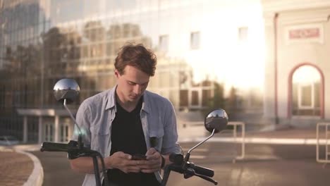 Outdoor-portrait-of-modern-young-man-with-mobile-phone-in-the-street,-sitting-on-minibike.-Modern-mirror-walls-building-on-the-background
