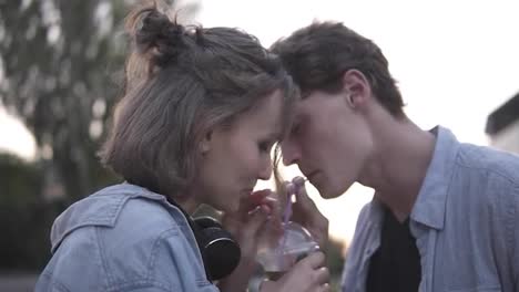 Young-attractive-couple-are-drinking-from-a-plastic-cup-together.-Standing-in-the-park-then-kissing-with-tenderness.-Slow-motion