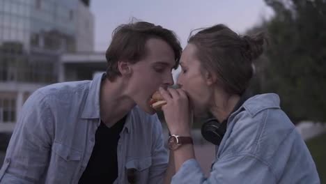 Beautiful-young-couple-enjoying-a-meal-together.-Eating-hamburger-together-from-the-sides.-Evening-dusk,-park-side