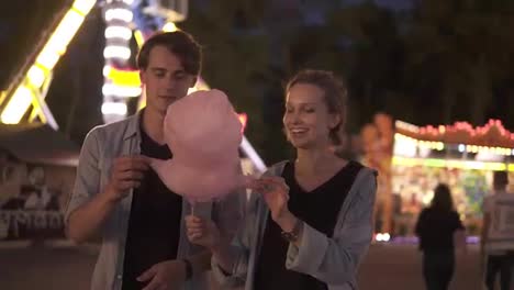 Young-hipster-couple-are-having-time-together-in-the-amusement-park-at-night.-Feeding-each-other-with-pink-cotton-candy.-Front-view