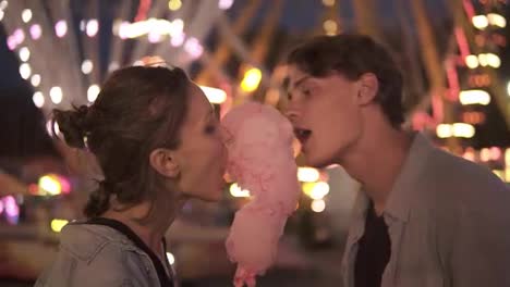 Lovely-young-couple-at-amusement-park.-Spending-time-together,-eating-pink-cotton-candy-from-sides.-Loving-couple,-dating.-Blurred-funfair-view-on-the-background