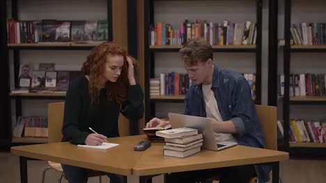 Young-male-and-female-students-talking,-explaining-course-work-study-together-in-library.-Actively-discussing-something-sitting-by-wooden-table-with-a-book.-Front-view