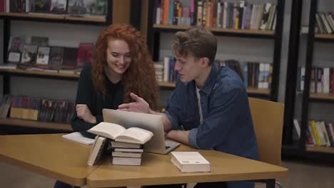 Young-male-and-female-students-talking-using-laptop-explaining-course-work-study-together-in-library.-Actively-discussing-something-sitting-by-wooden-table.-Slow-motion
