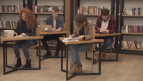 Group-of-students-preparing-for-exam-in-university-library,-two-beautiful-females-making-notes,-two-mates-behind-reading-book,-working-on-laptop,-concentrated-students-in-studying-process